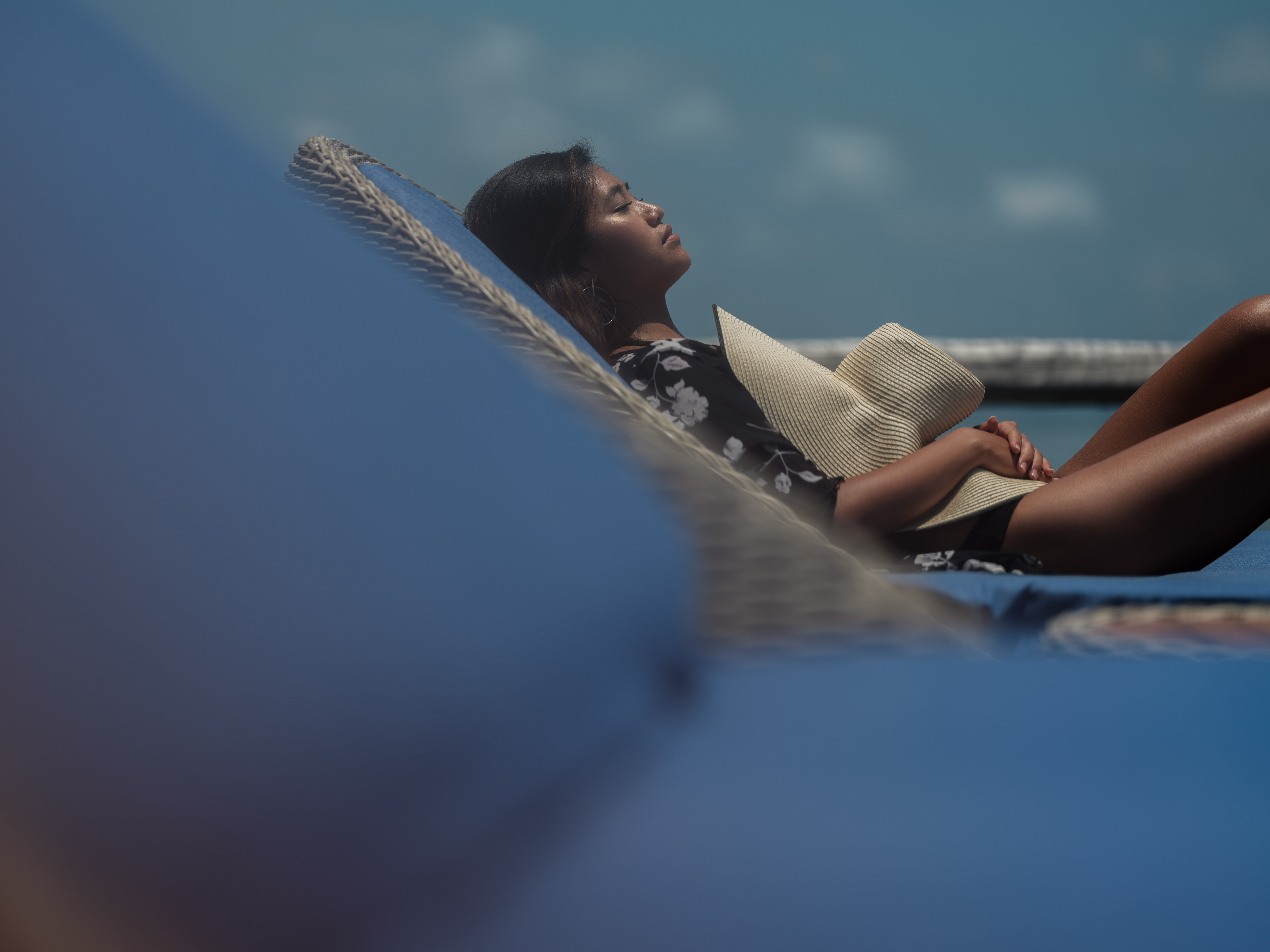 close_up_asian_girl_black_robe_holds_straw_hat_sun_bathing_on_blue_day_bed_overwater_suite