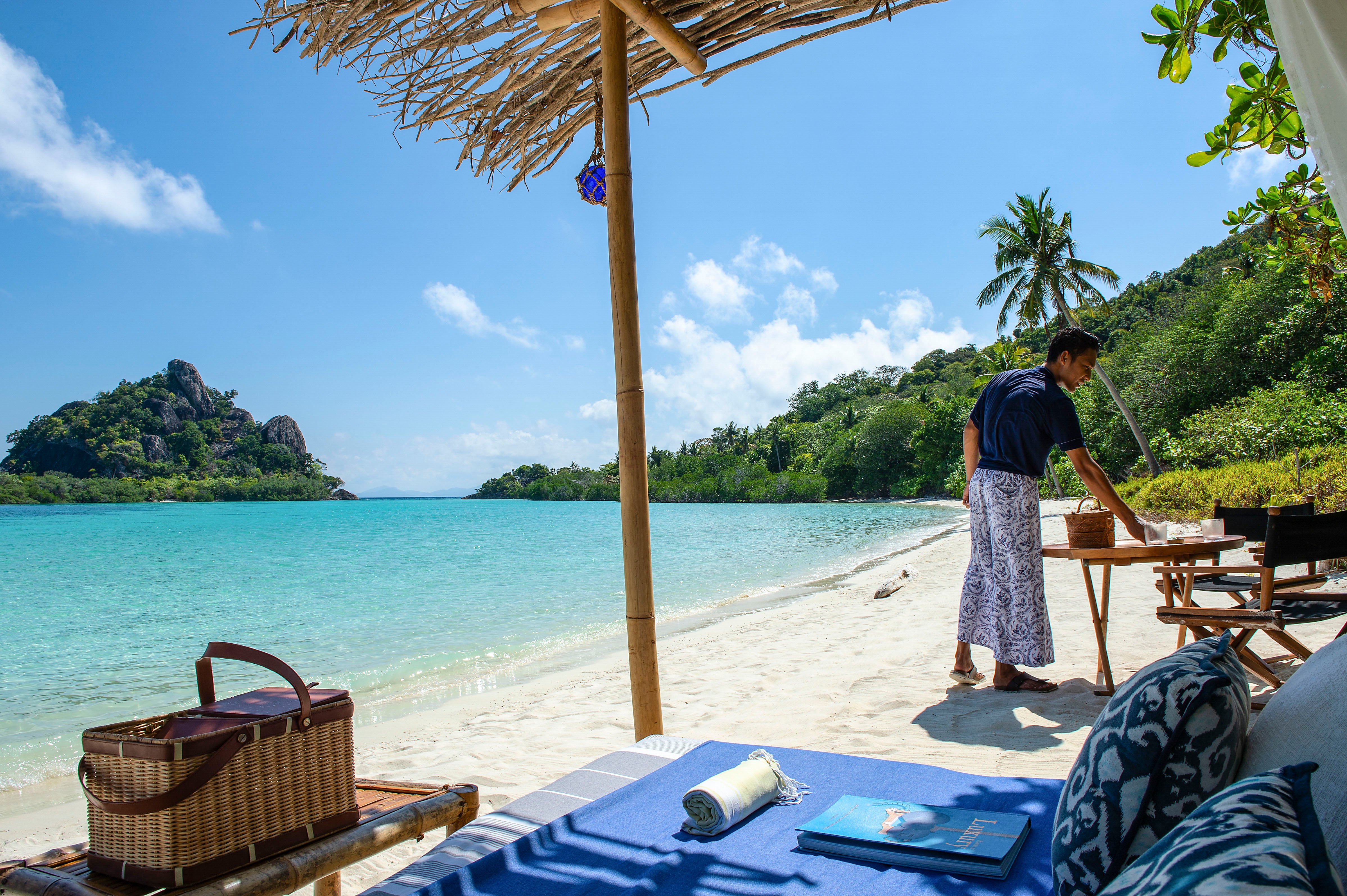 young_male_staff_batik_uniform_arranging_table_arm_chairs_coconut_beach_picnic_batu_tokong_background