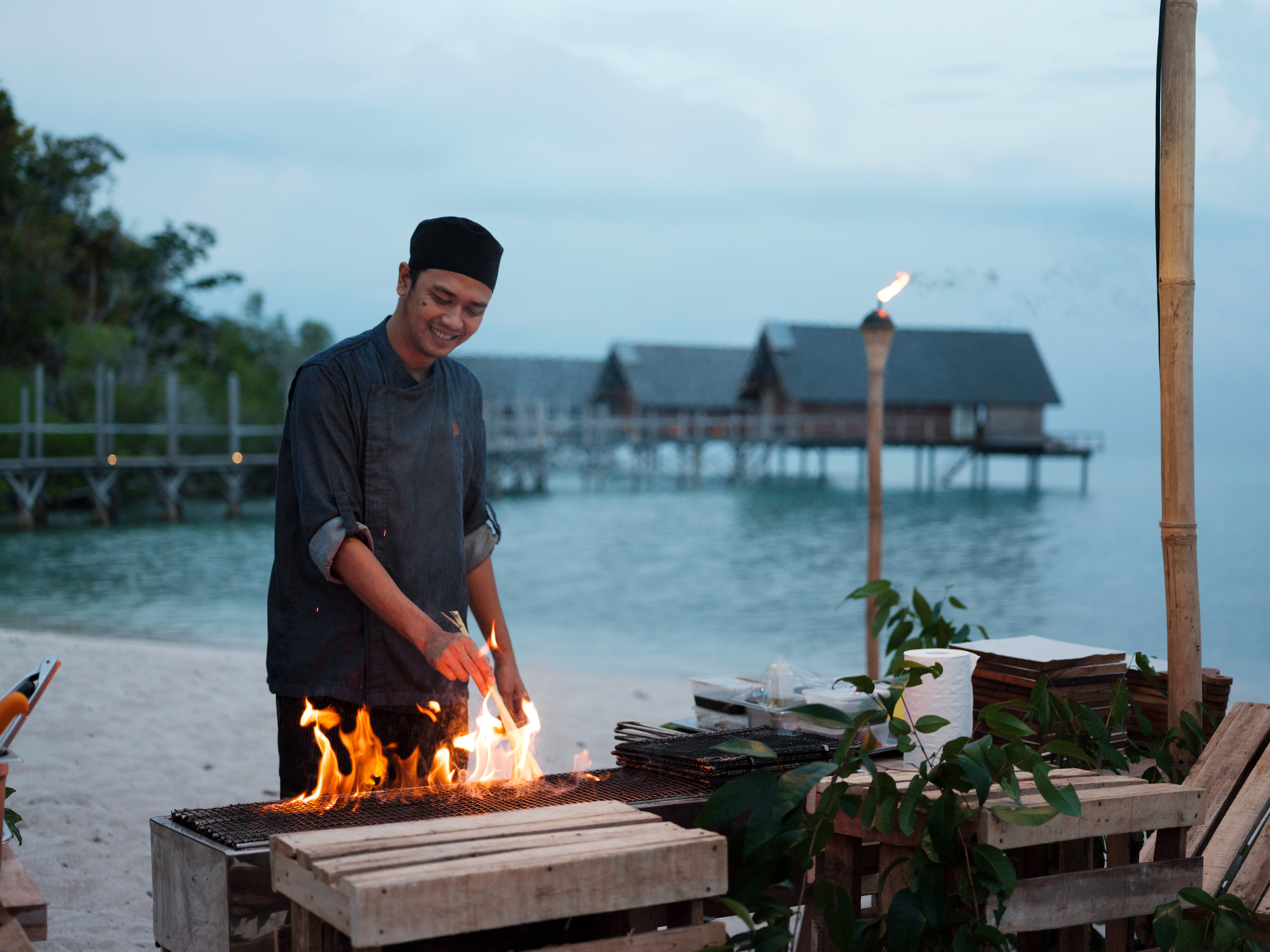 happy_male_chef_setting_barbeque_fire_in_beach_boat_house_east_overwater_suites_background