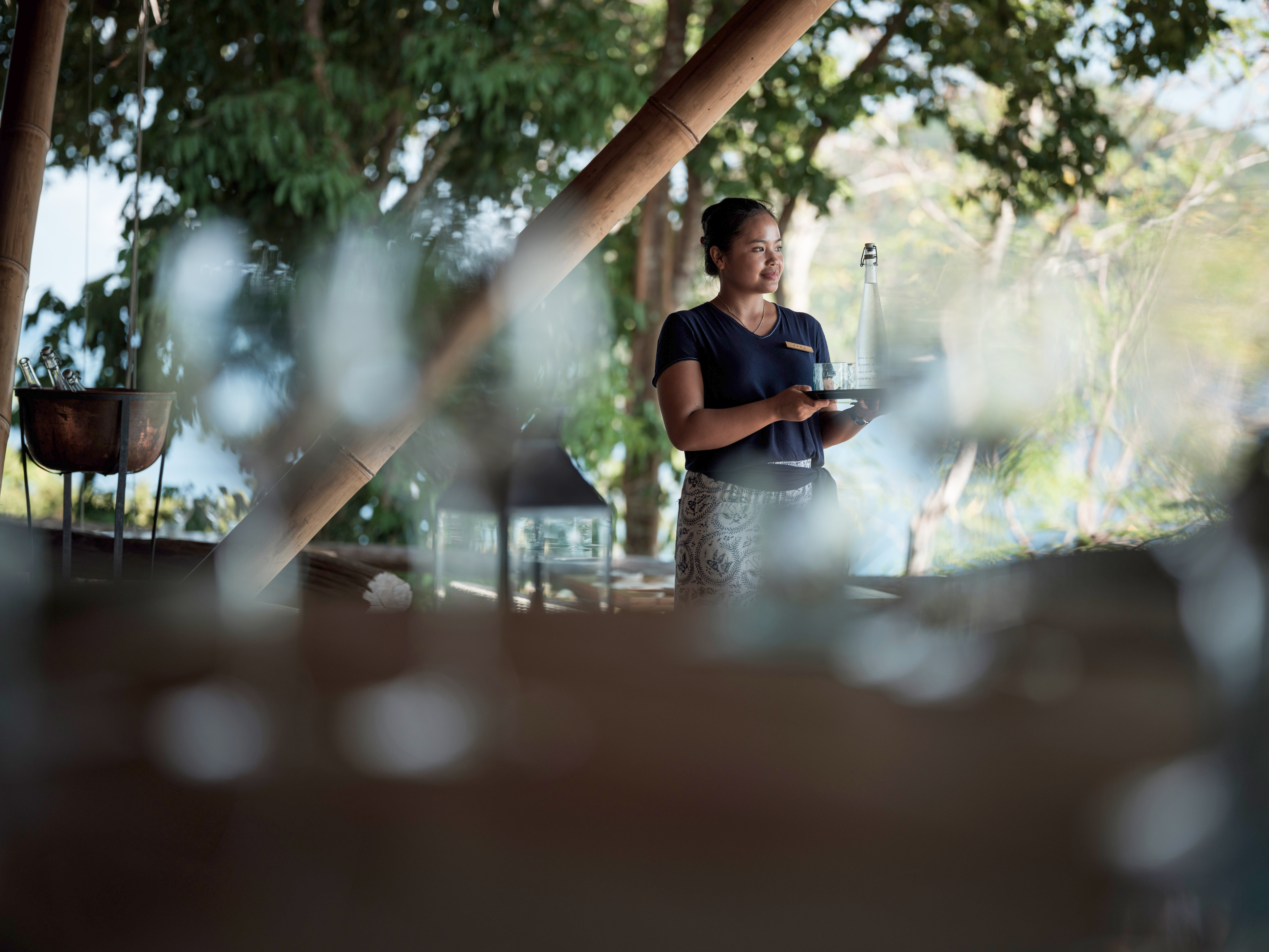female_staff_holding_glass_water_bottle_tray_treetops_deck_with_copper_ice_bucket_blurred_foliage_background