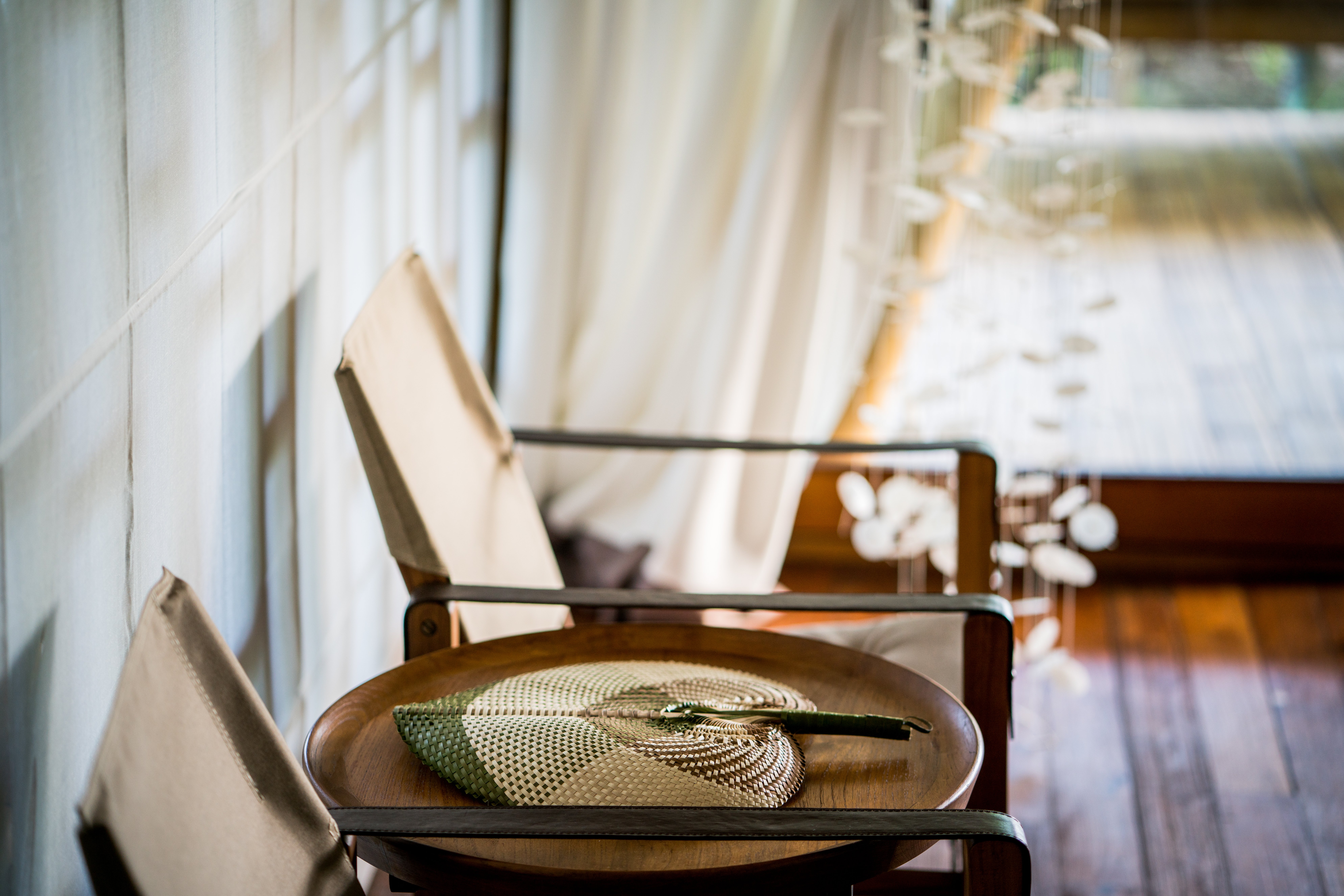 green_brown_rattan_hand_fan_on_wooden_table_arm_chairs_iwith_white_curtain_behind_blurred_jellyfish_tentacles_light_beach_suite