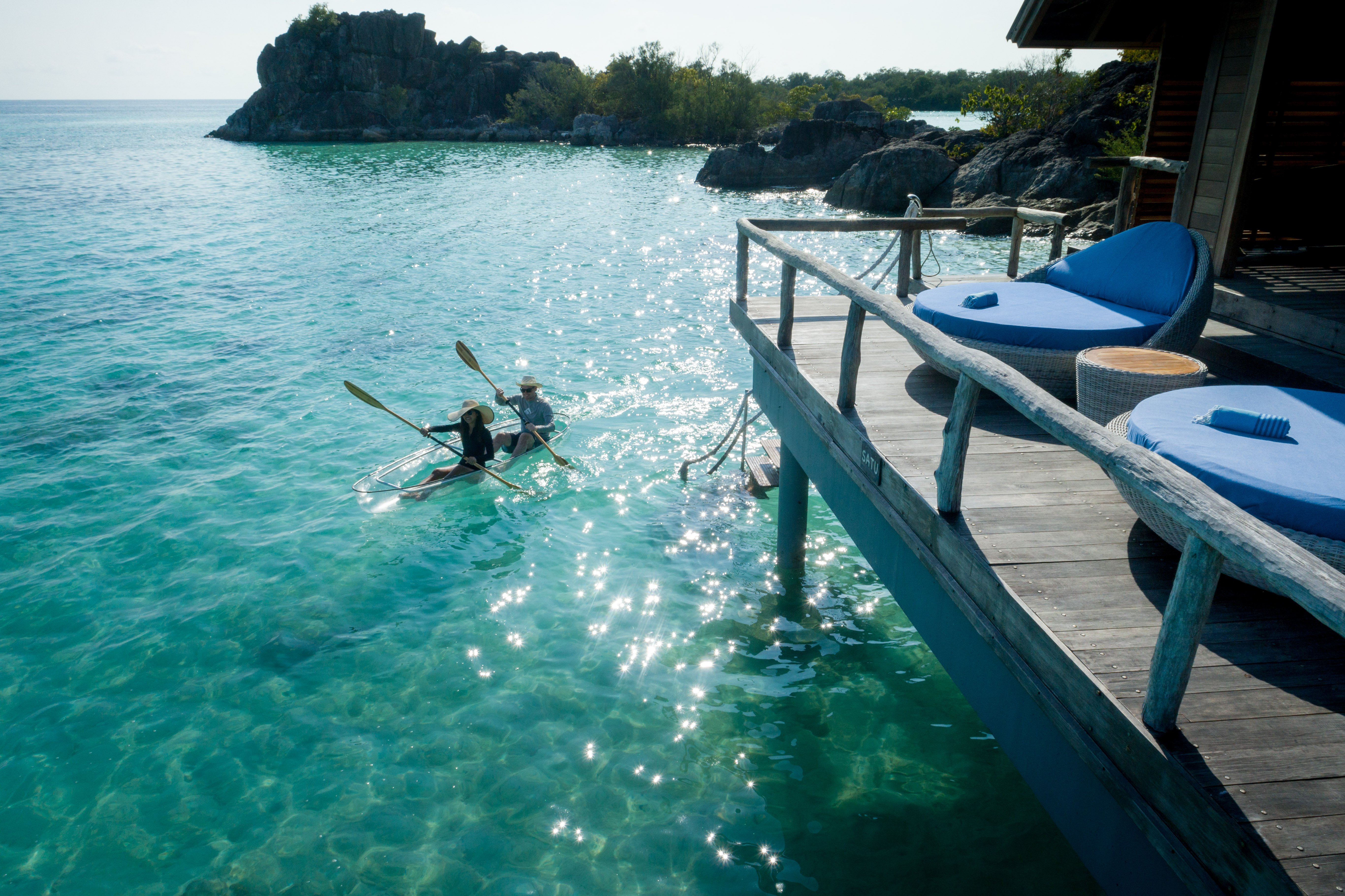 happy_couple_clear_kayaking_in_lagoon_under_overwater_suite_sun_terrace_blue_cushion_rattan_sun_beds_big_rocks_background