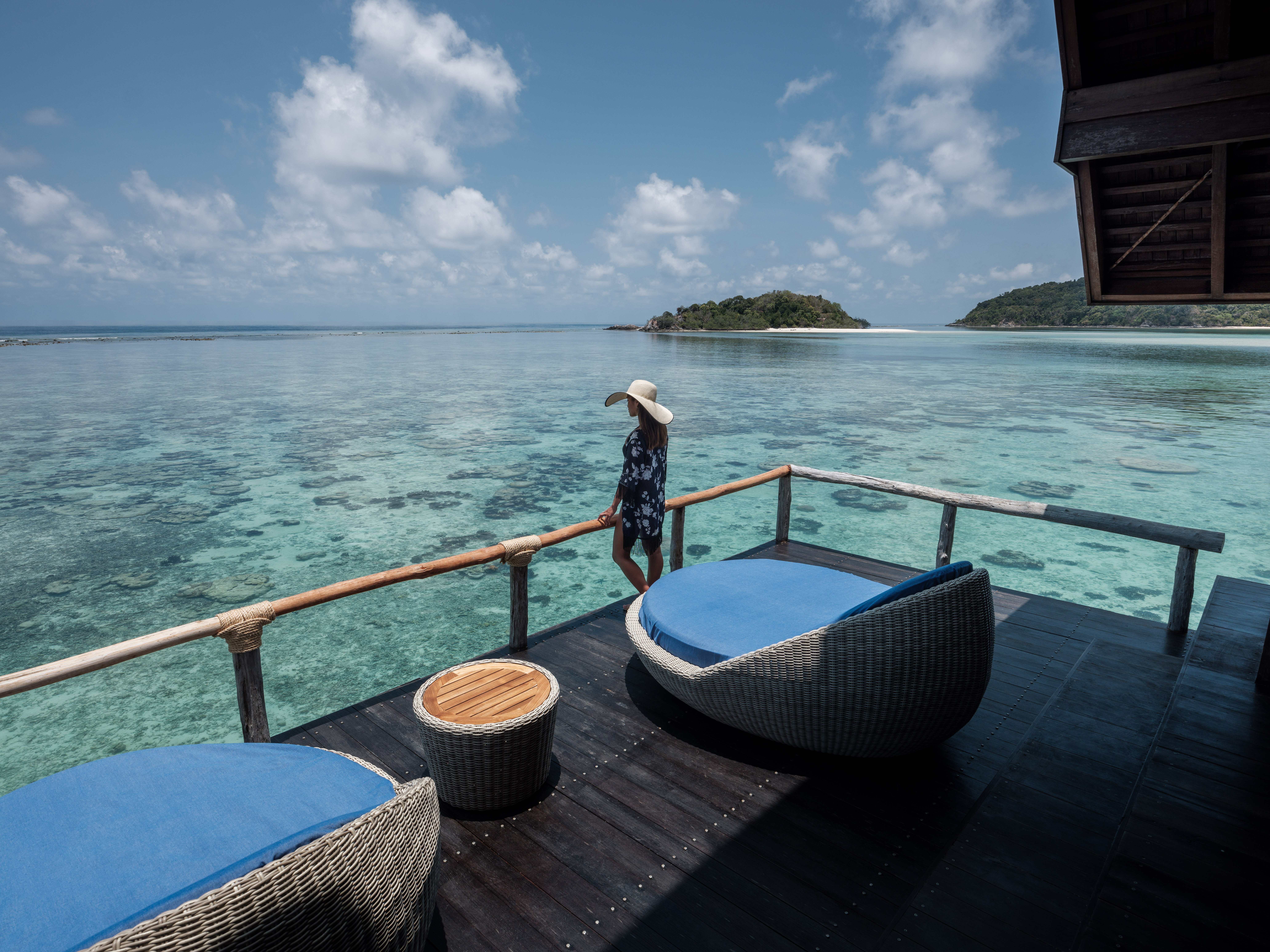 asian_girl_straw_hat_black_robe_hand_wood_banister_standing_overwater_sun_terrace_admiring_view_rattan_sun_beds_muerba_behind_with_sanggah_background