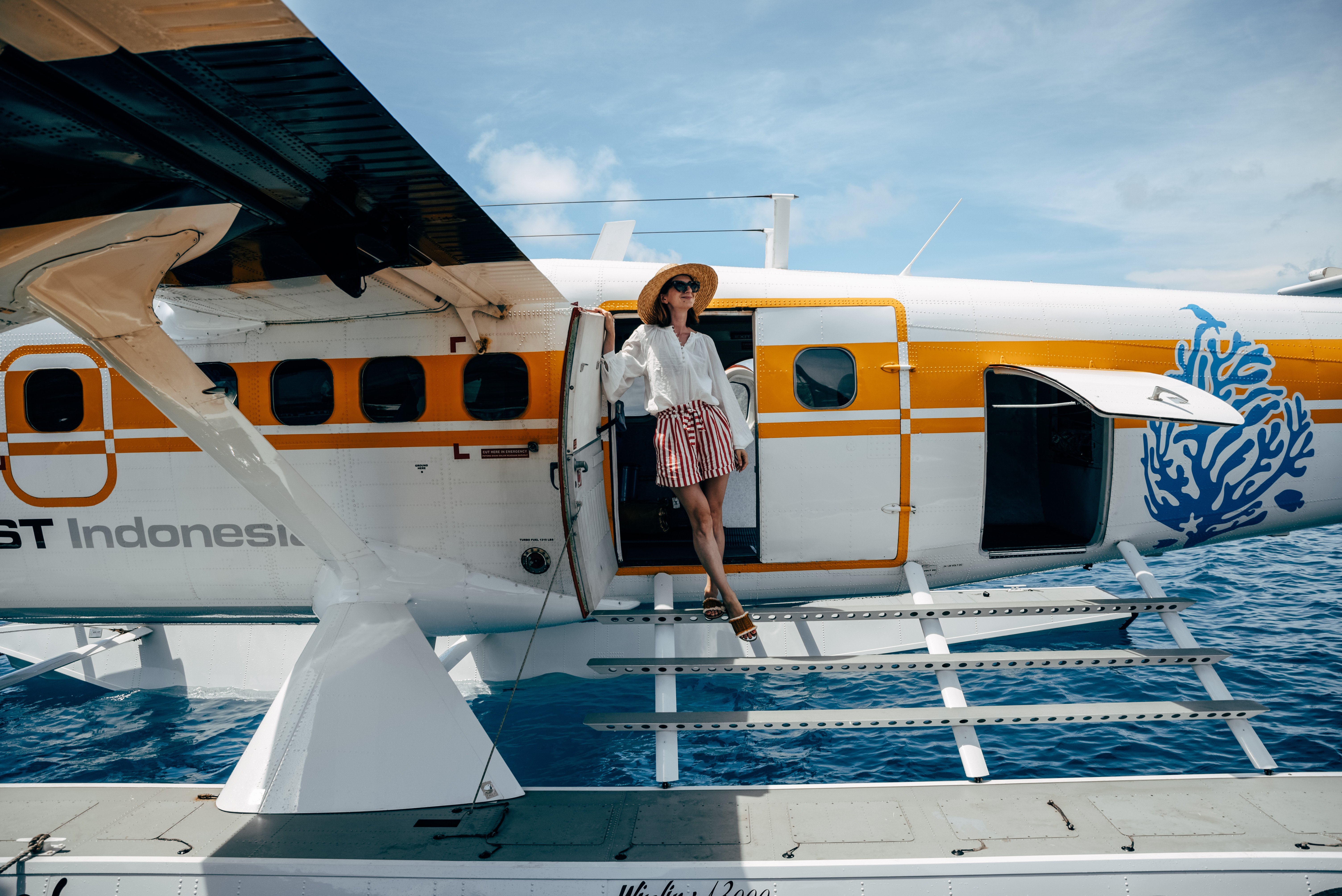 white_woman_sunglasses_straw_hat_striped_shorts_standing_in_front_of_bawah_seaplane_stairs