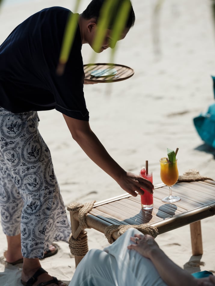 young_male_staff_batik_uniform_placing_juice_on_bamboo_table_in_beach